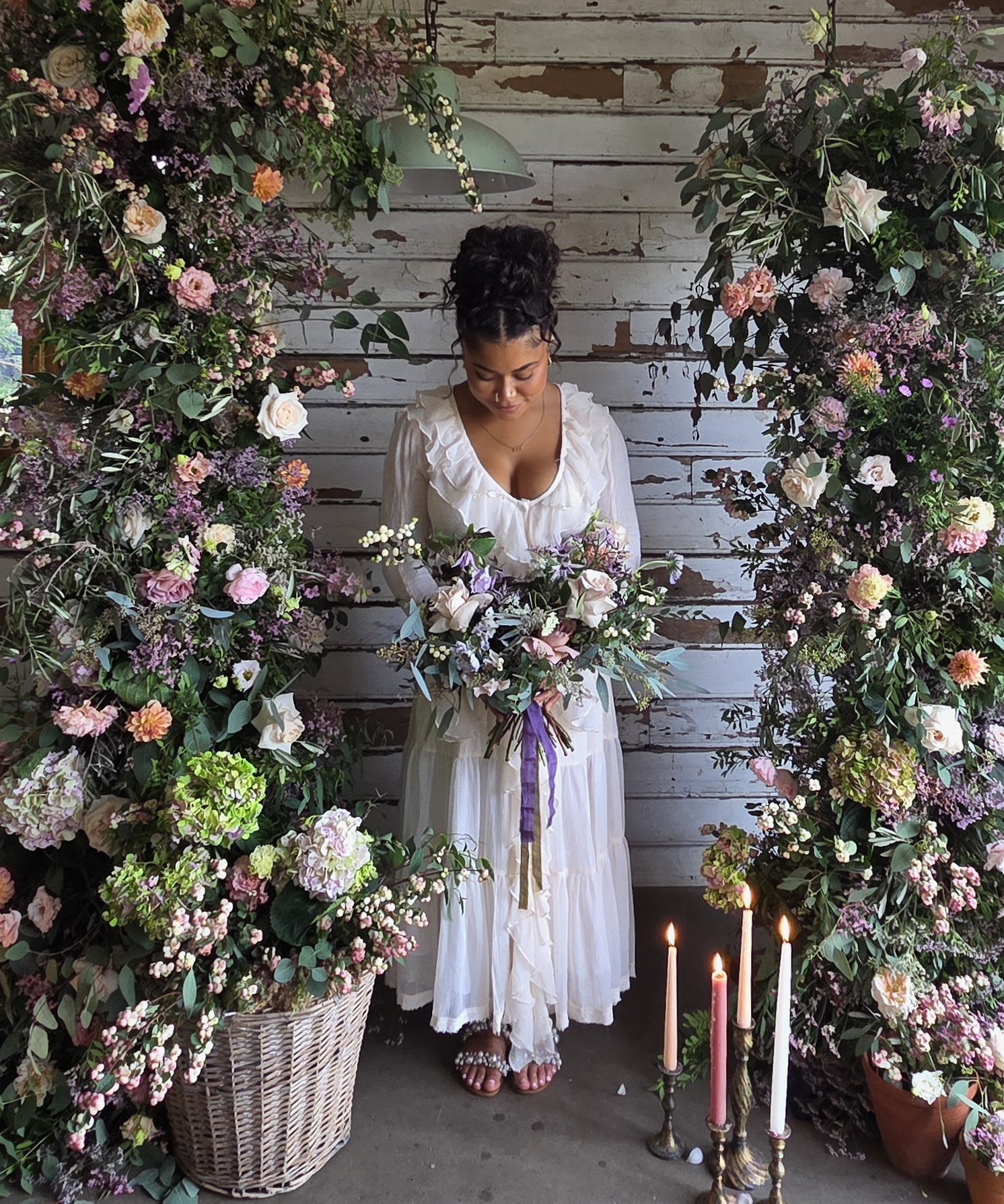 A woman dressed in a white bohemian-style dress stands amidst an arch of pink, purple, and white flowers in front of a weathered white wooden wall. Candles in a metal candelabrum are lit in front of her, and a large basket of flowers is on her left.