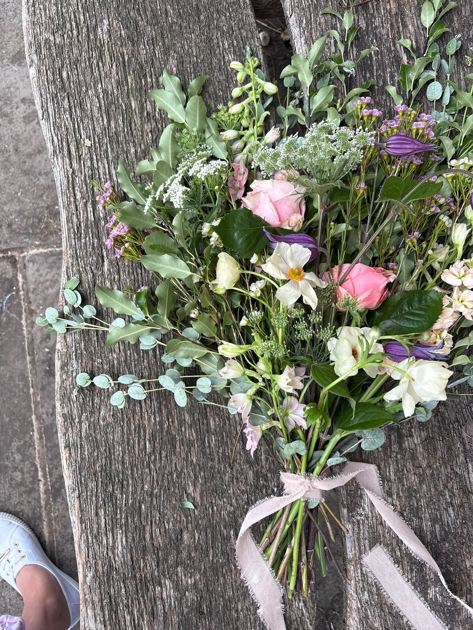 A bouquet of mixed flowers with pink, white, and purple blooms and greenery, tied with a light pink ribbon, resting on a weathered wooden surface.