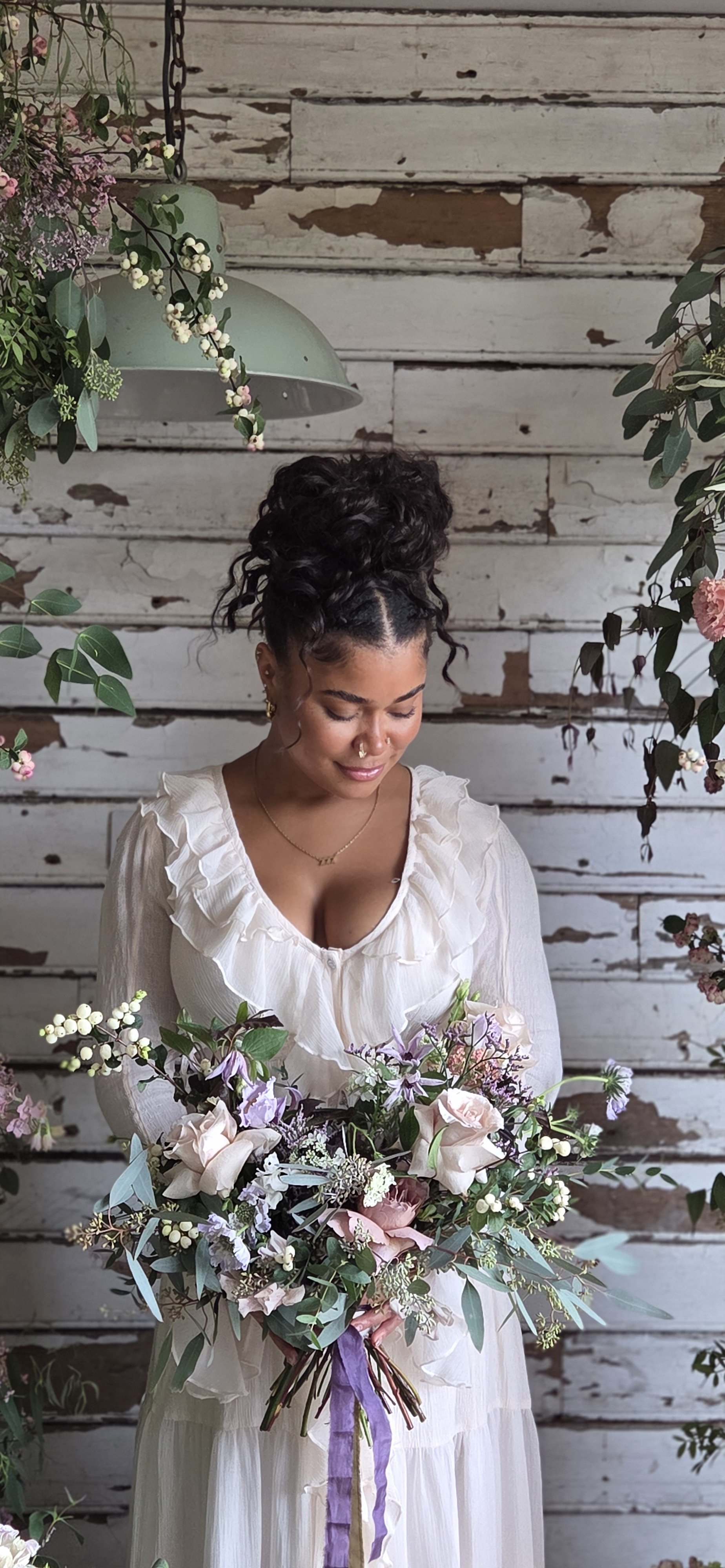 A woman holding a large bouquet of flowers, standing in front of a rustic wooden wall decorated with hanging greenery and flowers, dressed in a cream-colored ruffled dress.