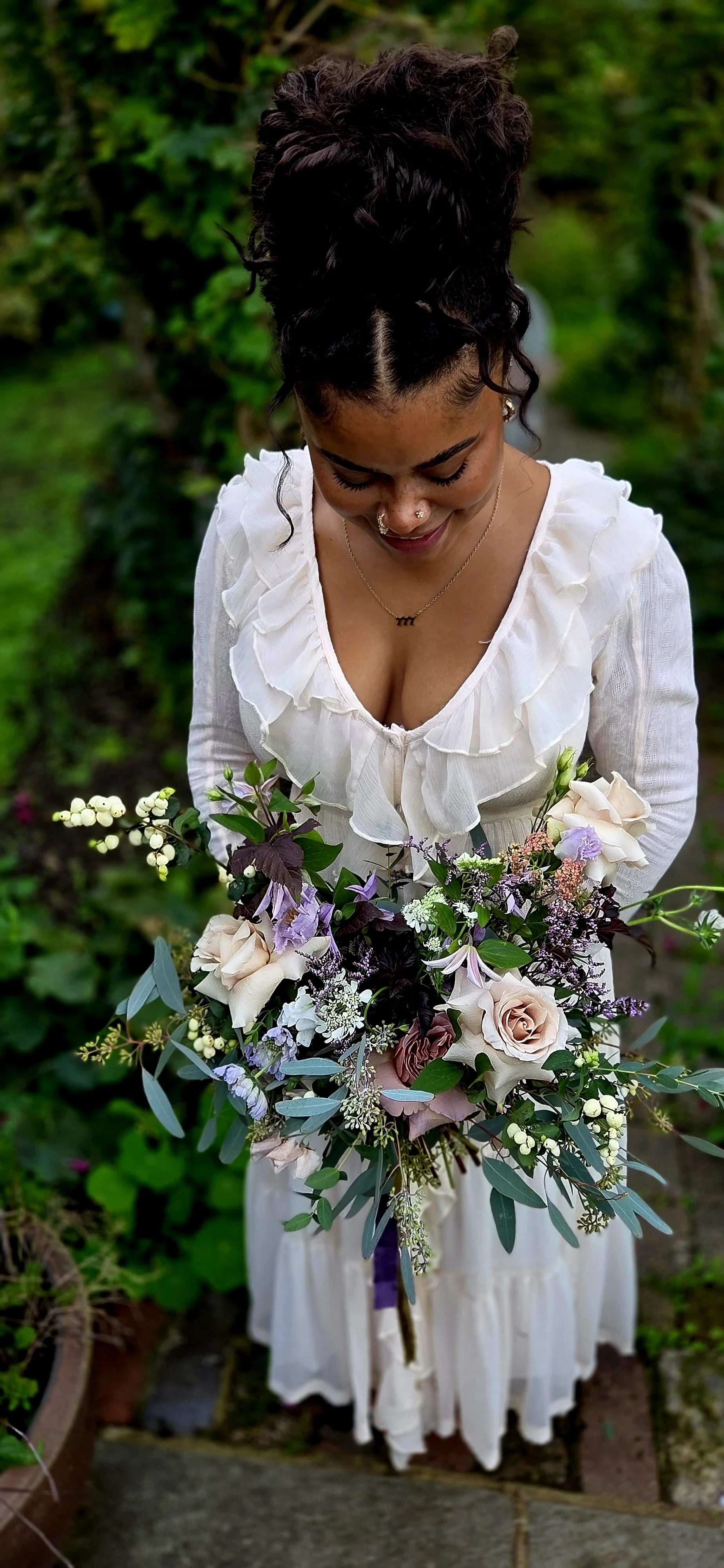A woman wearing a white dress with ruffled neckline, holding a large bouquet of flowers including roses and lavender, standing outdoors with greenery in the background.