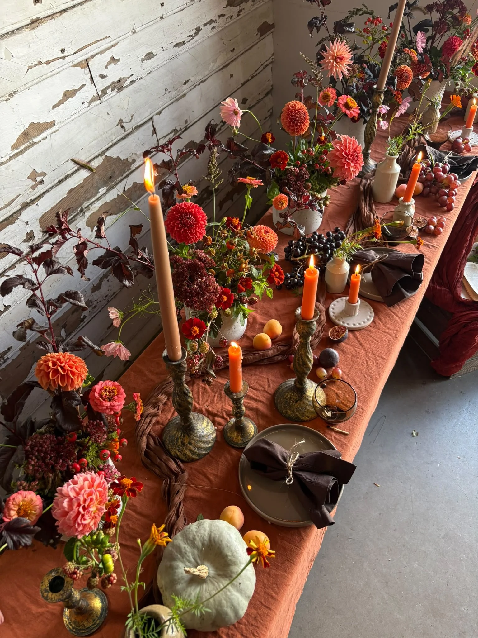 A long rustic table decorated with a variety of vibrant flowers, candles, grapes, pumpkins, and tableware, set against a distressed white wooden wall.