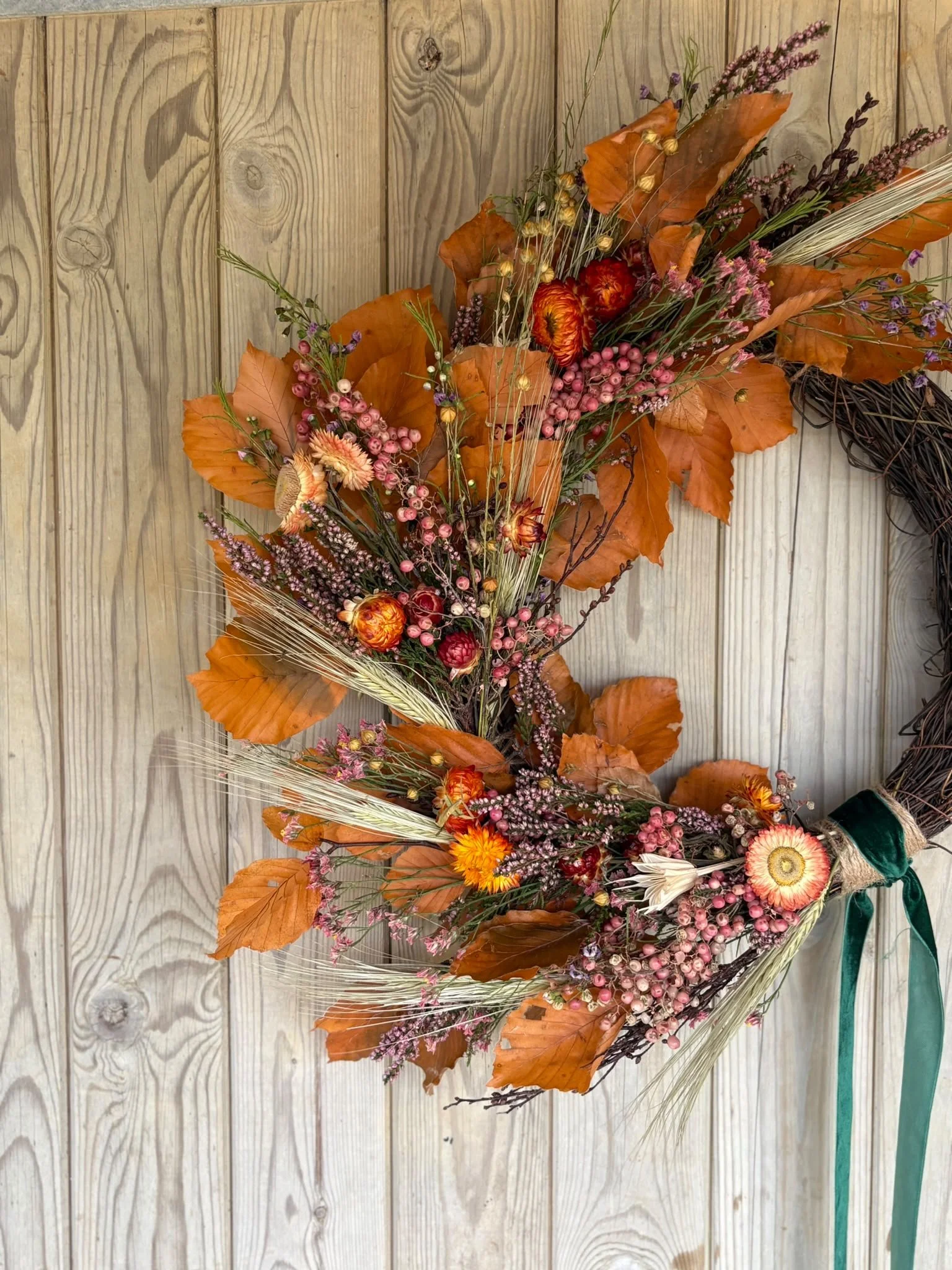 A dried flower and leaf arrangement on a wooden surface, featuring orange leaves, pink berries, small pink flowers, and grasses.