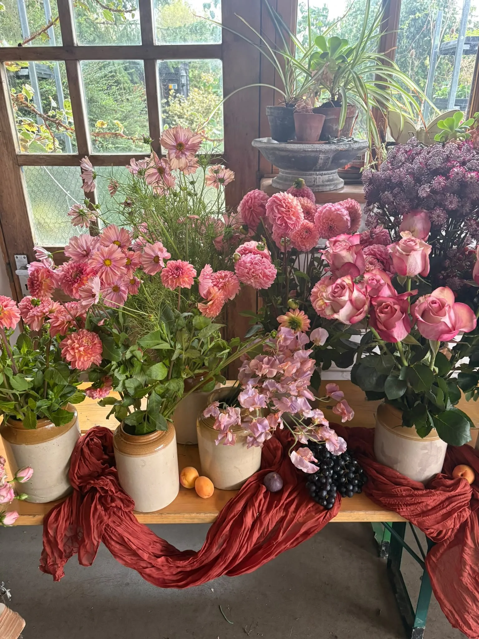 An indoor display of pink and purple flowers in ceramic vases on a wooden table, with a backdrop of a window and green outdoor foliage, decorated with red fabric.