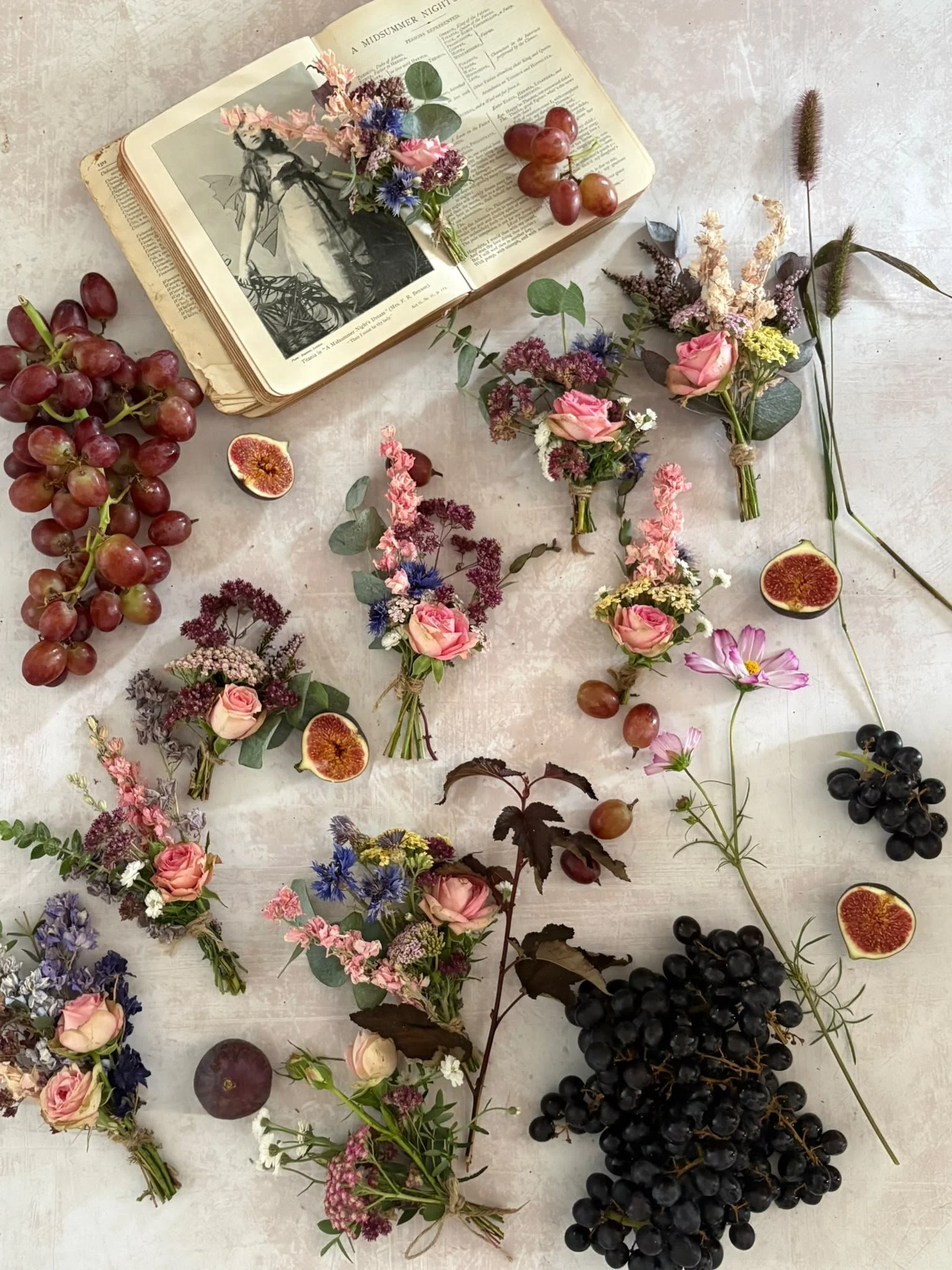 A flat lay of various flowers, grapes, and an open book on a light-colored surface. The flowers are in small bouquets, some tied with twine, and include roses, lavender, and other wildflowers. There are clusters of red and black grapes, some halved f