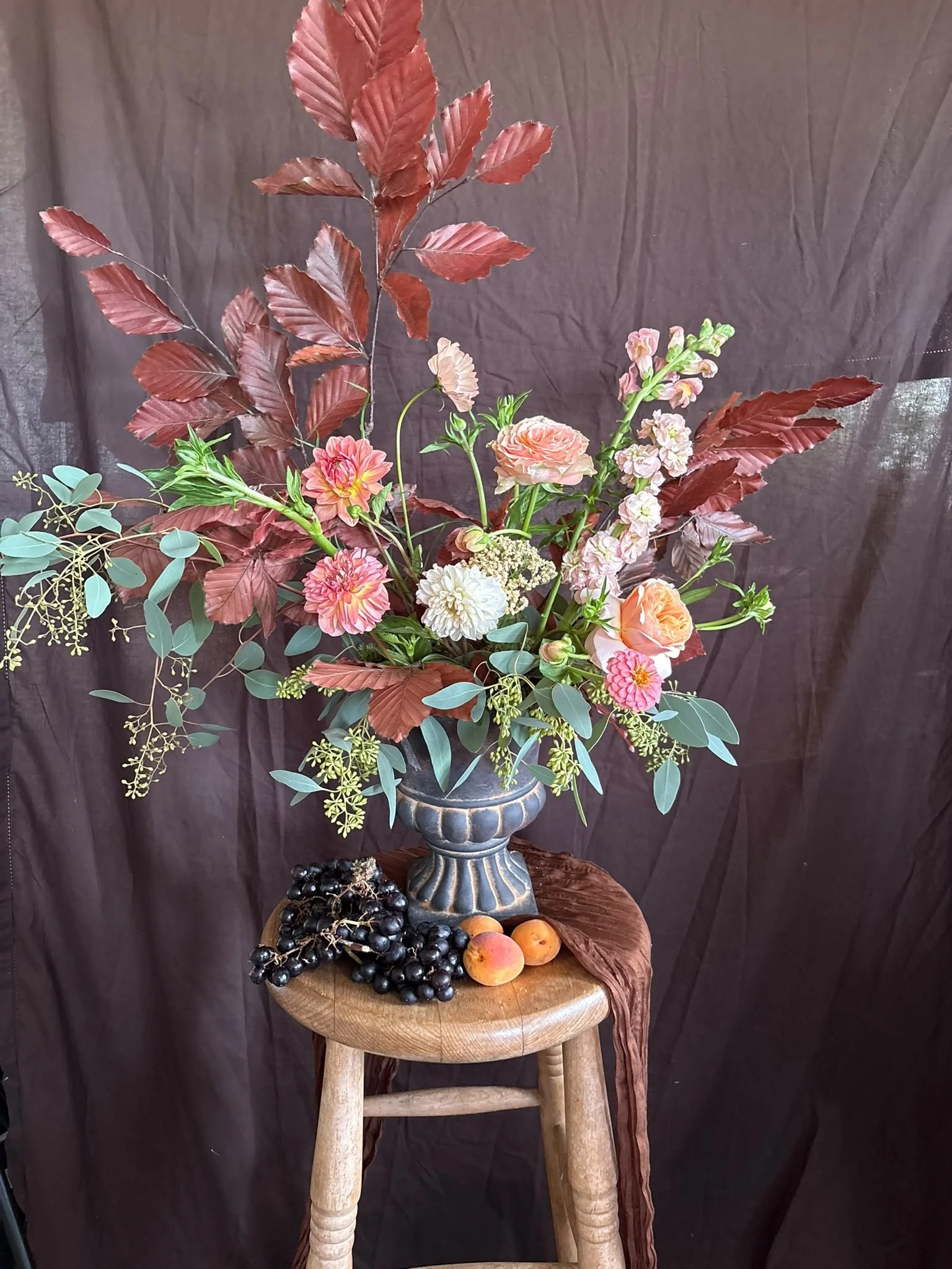 A floral arrangement of pink, white, and peach flowers with green leaves and large reddish-brown leaves in a silver vase on a wooden table, with dark background.