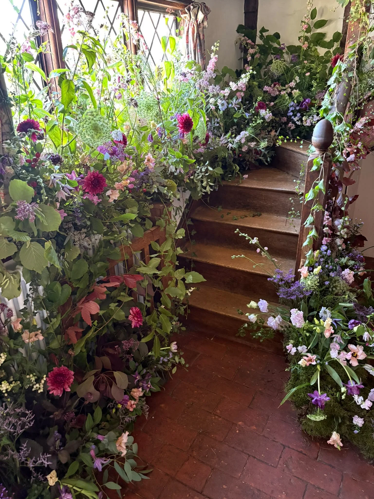 A wooden staircase surrounded by an abundance of flowering plants and greenery on both sides, with a window and curtains in the background.