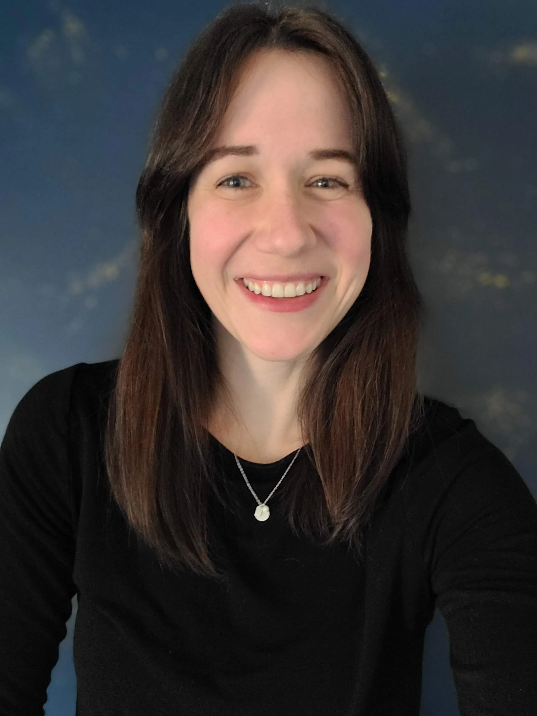 A woman with long brown hair wearing a black top, silver necklace with a pendant, and smiling in front of a dark blue background with faint clouds.