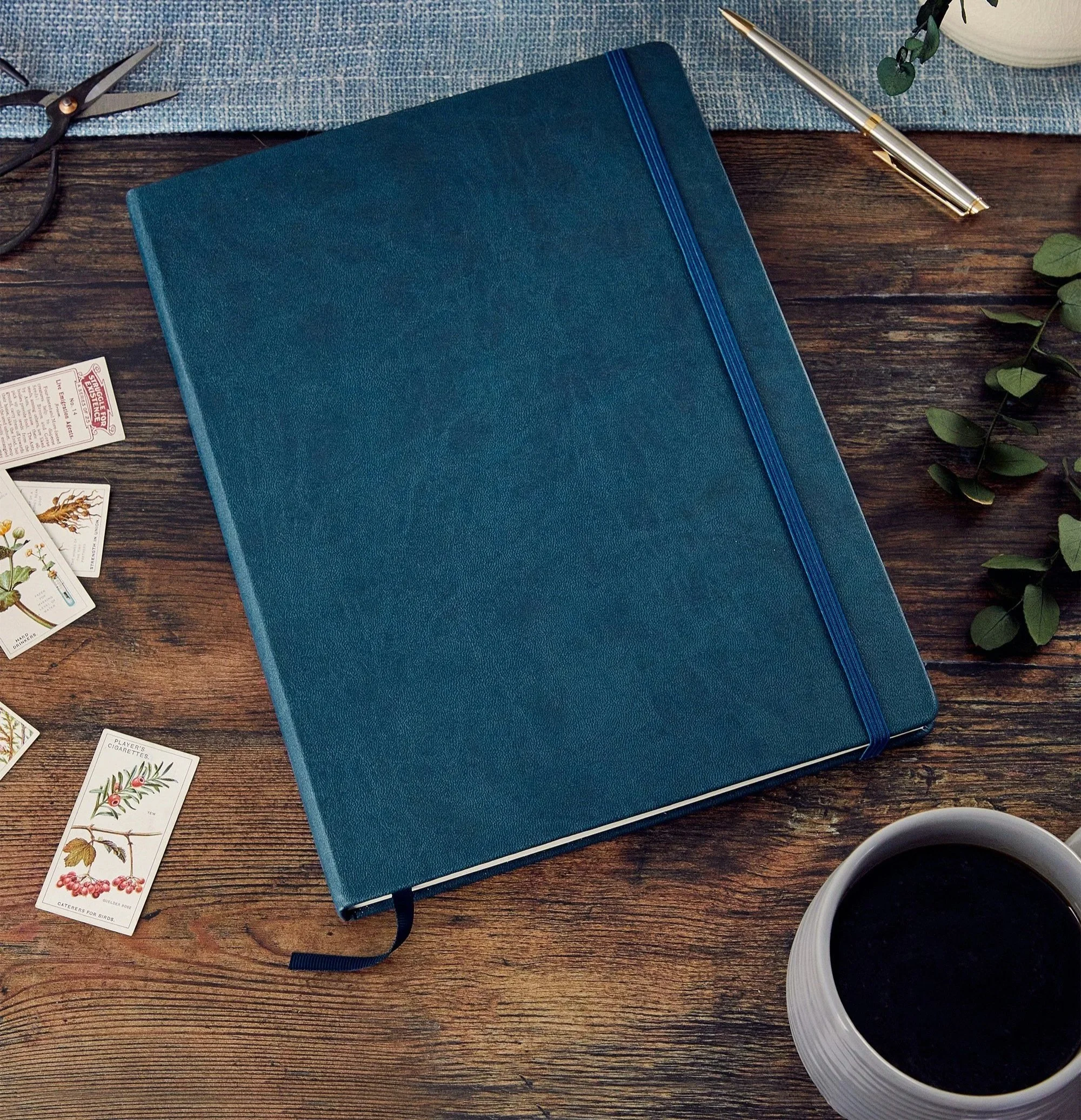 A blue notebook with black elastic band and two blue ribbons on a wooden table, alongside a gold pen, a pair of black scissors, gardening plant tags with botanical illustrations, a white cup of coffee, and some green leaves.