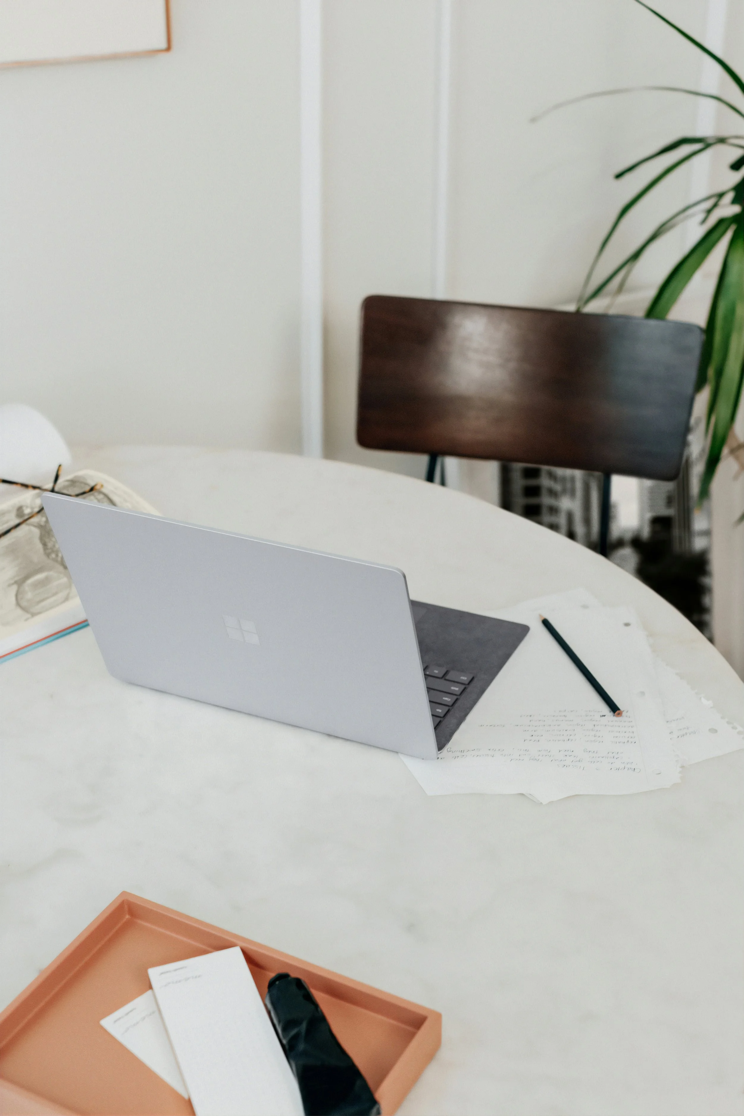 Office workspace with a white table, a Microsoft Surface laptop, papers, a black stapler, and a terracotta-colored tray with papers and a stapler inside. In the background, there is a wooden chair and a large plant in a pot.