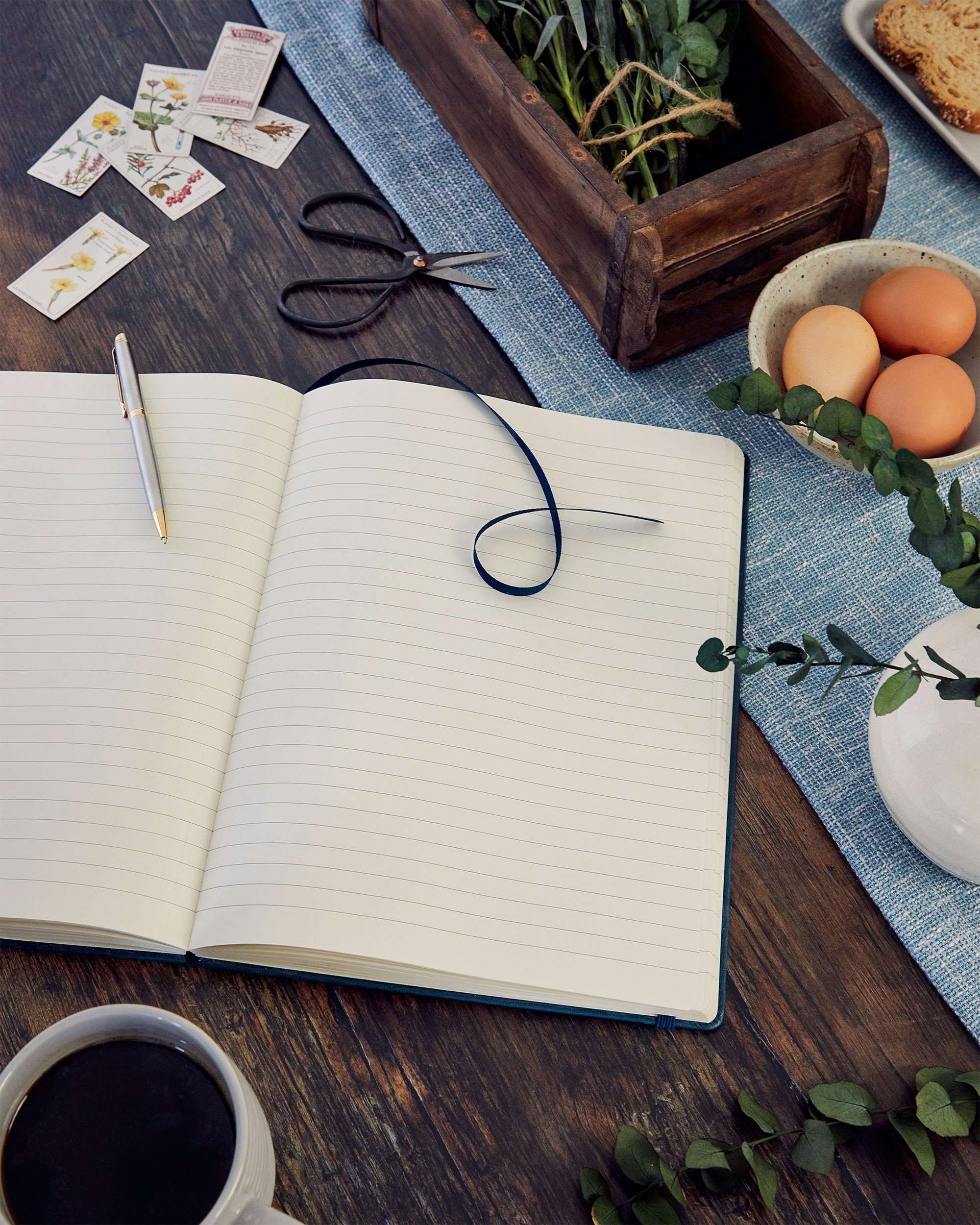 A wooden table with a blue cloth runner, a notebook with a pen, a cup of black coffee, a pair of scissors, seed packets, a ceramic bowl with three eggs, a green leafy plant in a wooden box, and a plate with two cookies.