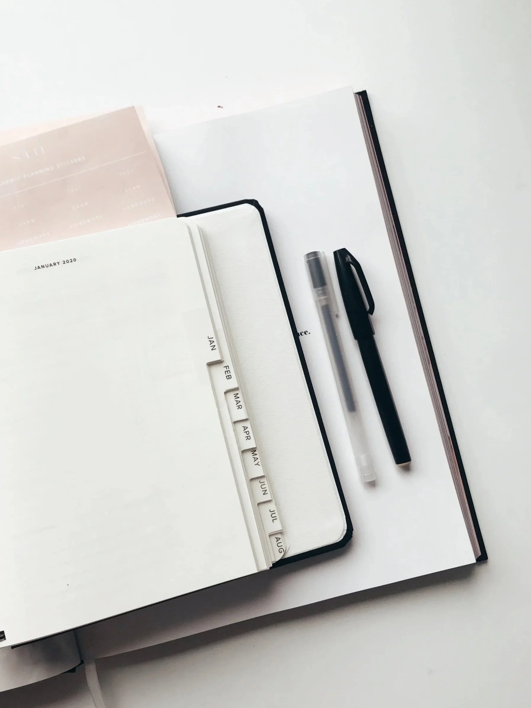 Open planner or journal with tabbed monthly divider, a black pen, and a white highlighter on a white surface.