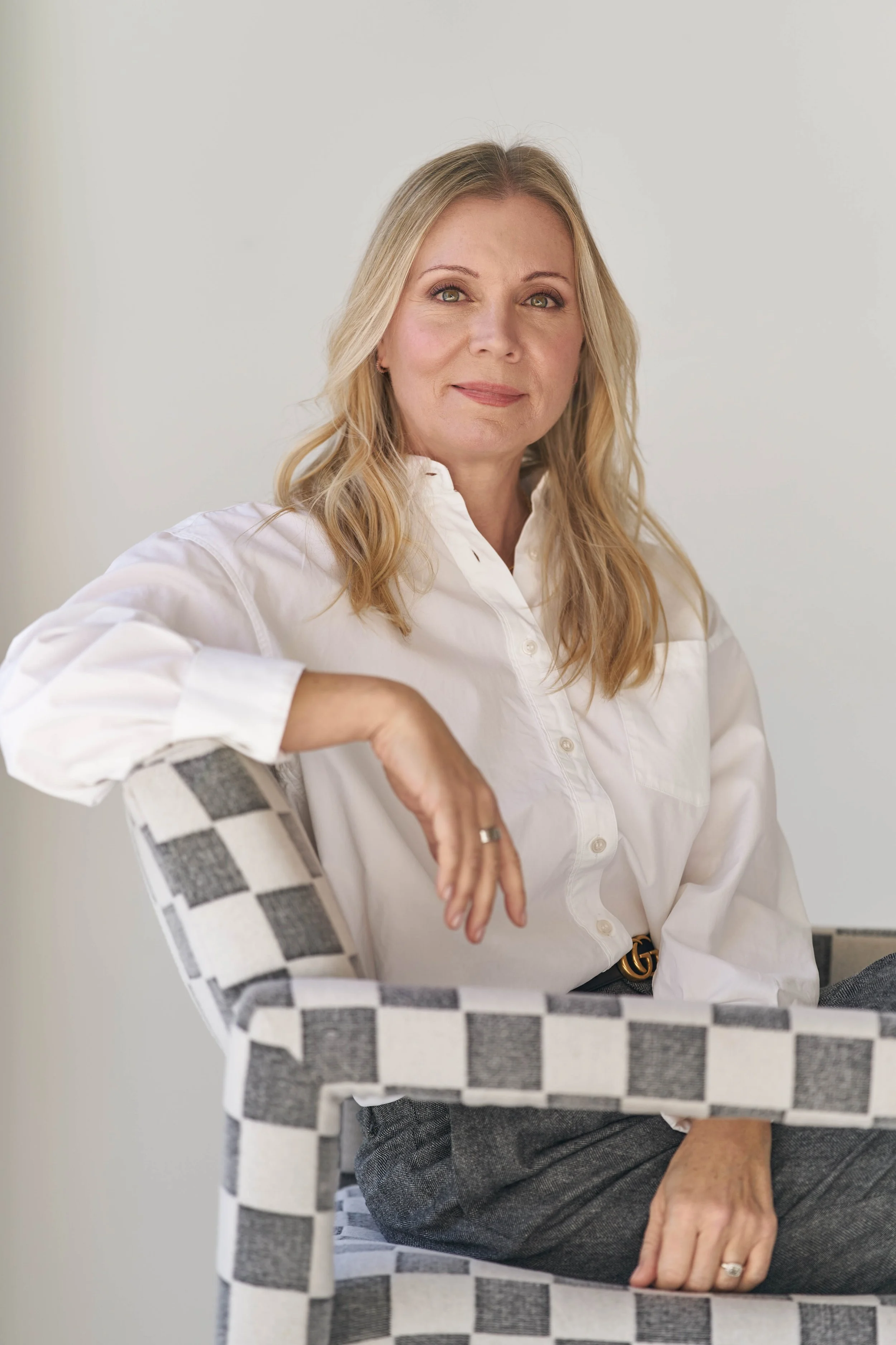 A woman with blonde hair and light green eyes, wearing a white shirt, sitting on a checkered chair with her arm resting on the back of the chair, against a plain light gray background.
