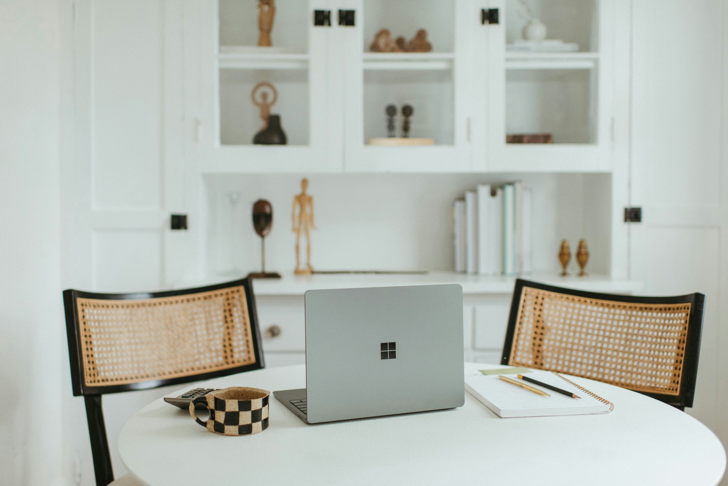 A white table with a Microsoft Surface laptop, a checkered mug, a notebook, and a pen. Two chairs with woven cane backs are visible in front of the table. A white cabinet with decorative items and books is in the background.