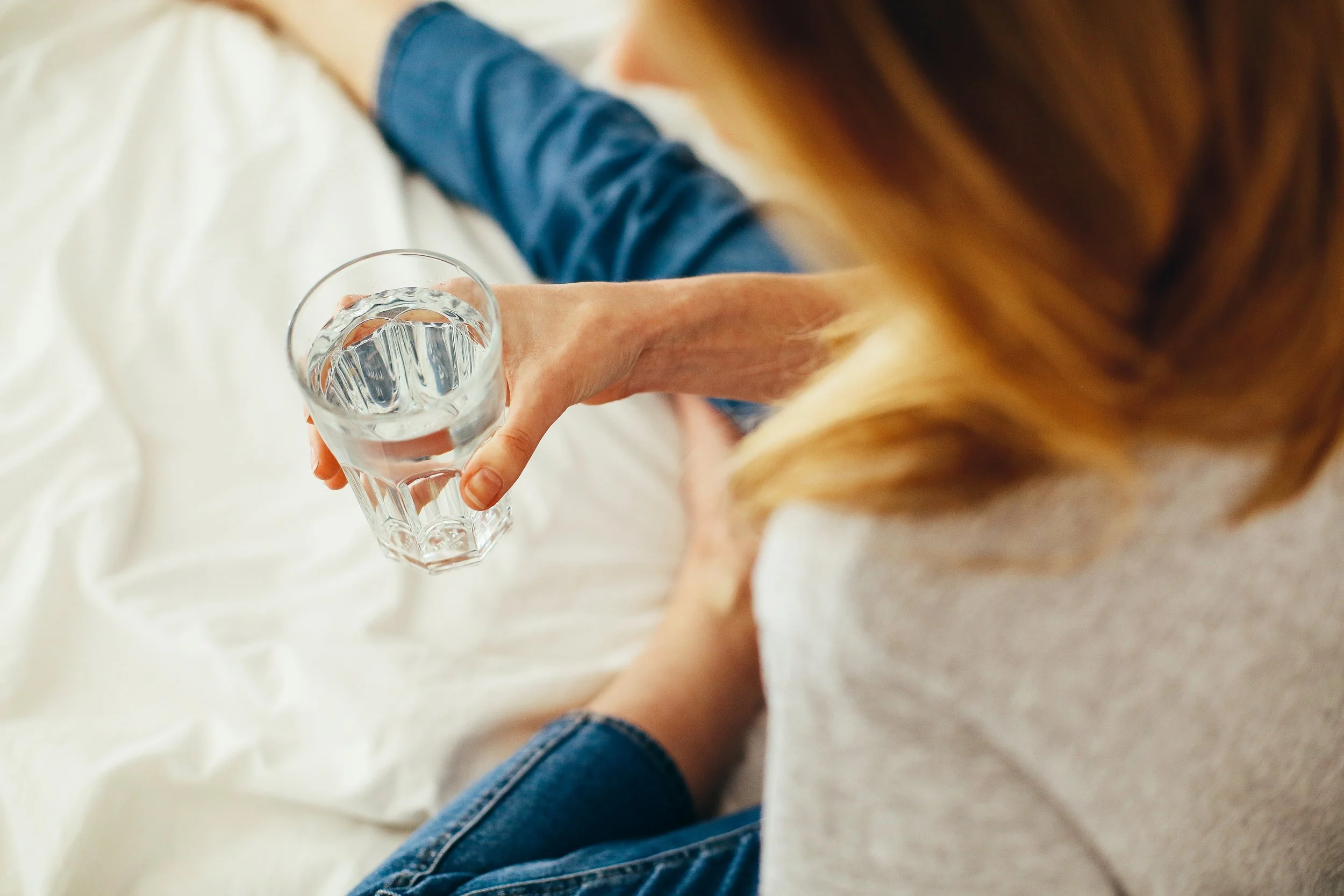 A person with long hair is sitting on a white surface, holding a glass of water. They are wearing a blue denim jacket and blue jeans.