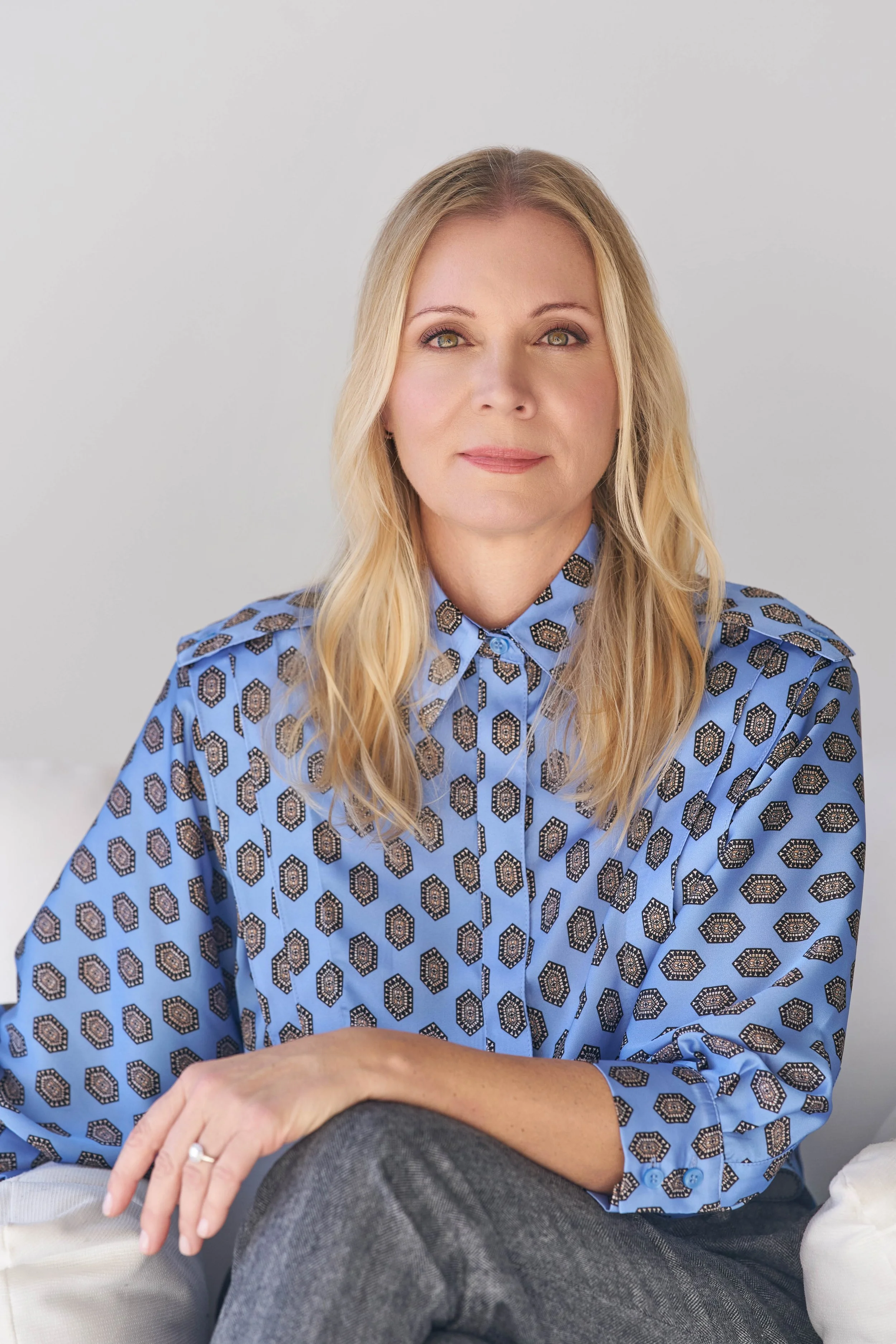 A woman with blonde hair and green eyes, wearing a blue patterned shirt, sits with her hand resting on her knee, in front of a plain white wall.
