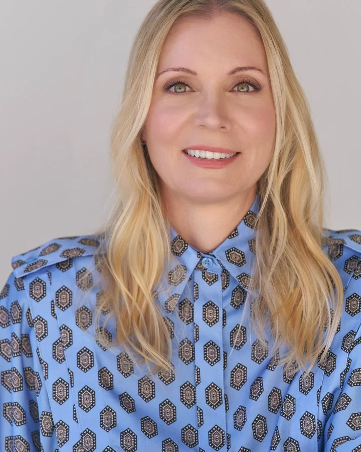 A woman with long blonde hair wearing a blue patterned shirt with geometric designs, smiling at the camera.