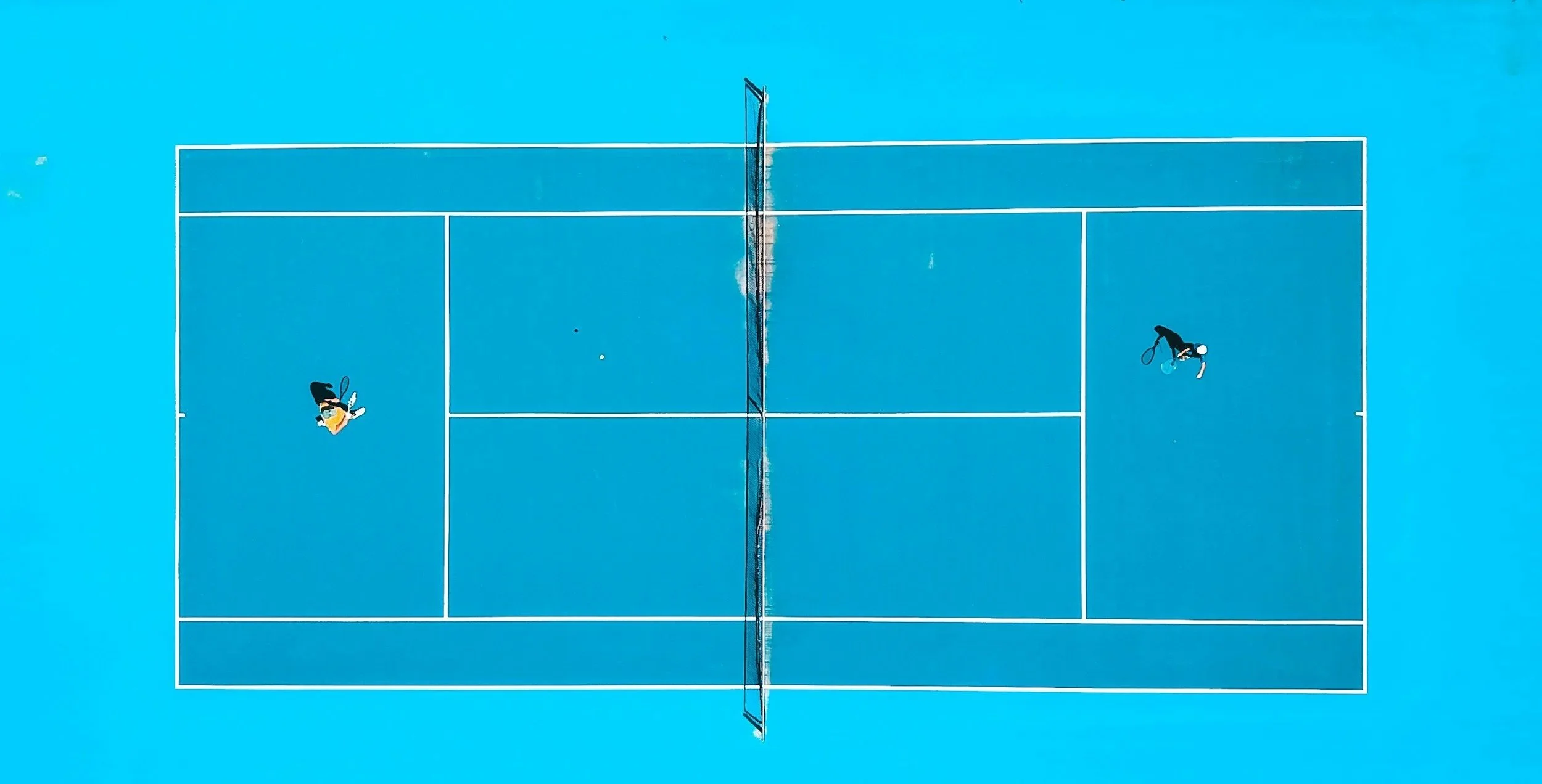 An aerial view of a blue tennis court with two players, one on each side, viewed from above.
