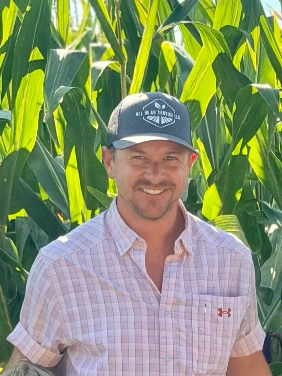 A man smiling outdoors in front of tall green corn plants, wearing a light pink checkered short-sleeve shirt and a gray cap with a logo and the text 'All in Ag Service LLC'.