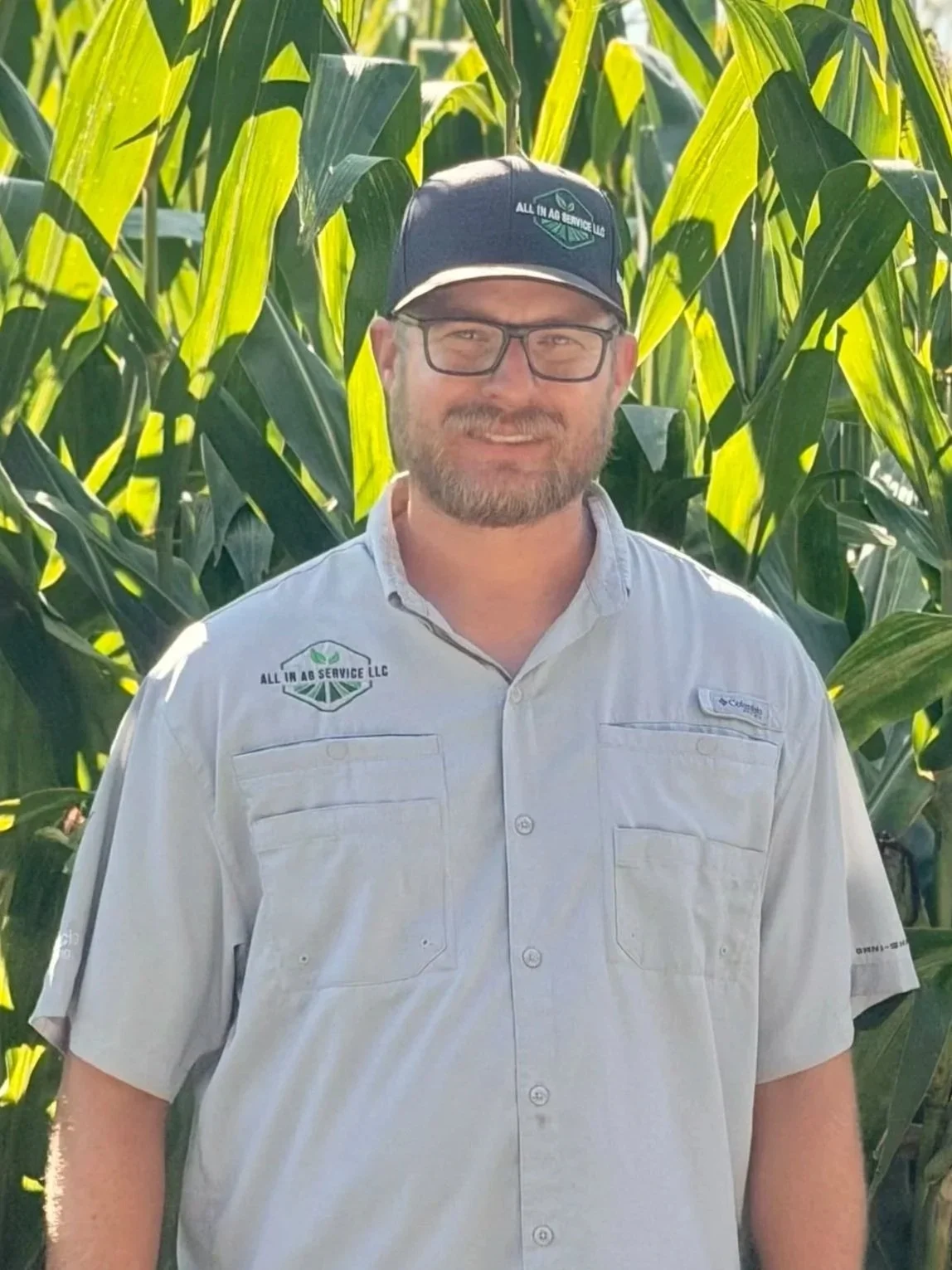 A man with glasses and a beard standing in front of tall green corn plants, wearing a light gray button-up shirt and a dark cap with the logo of All In Ag Service LLC.