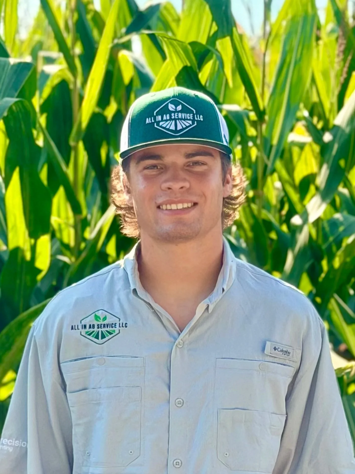 A man wearing a green cap and a light-colored shirt standing in front of tall green corn plants.