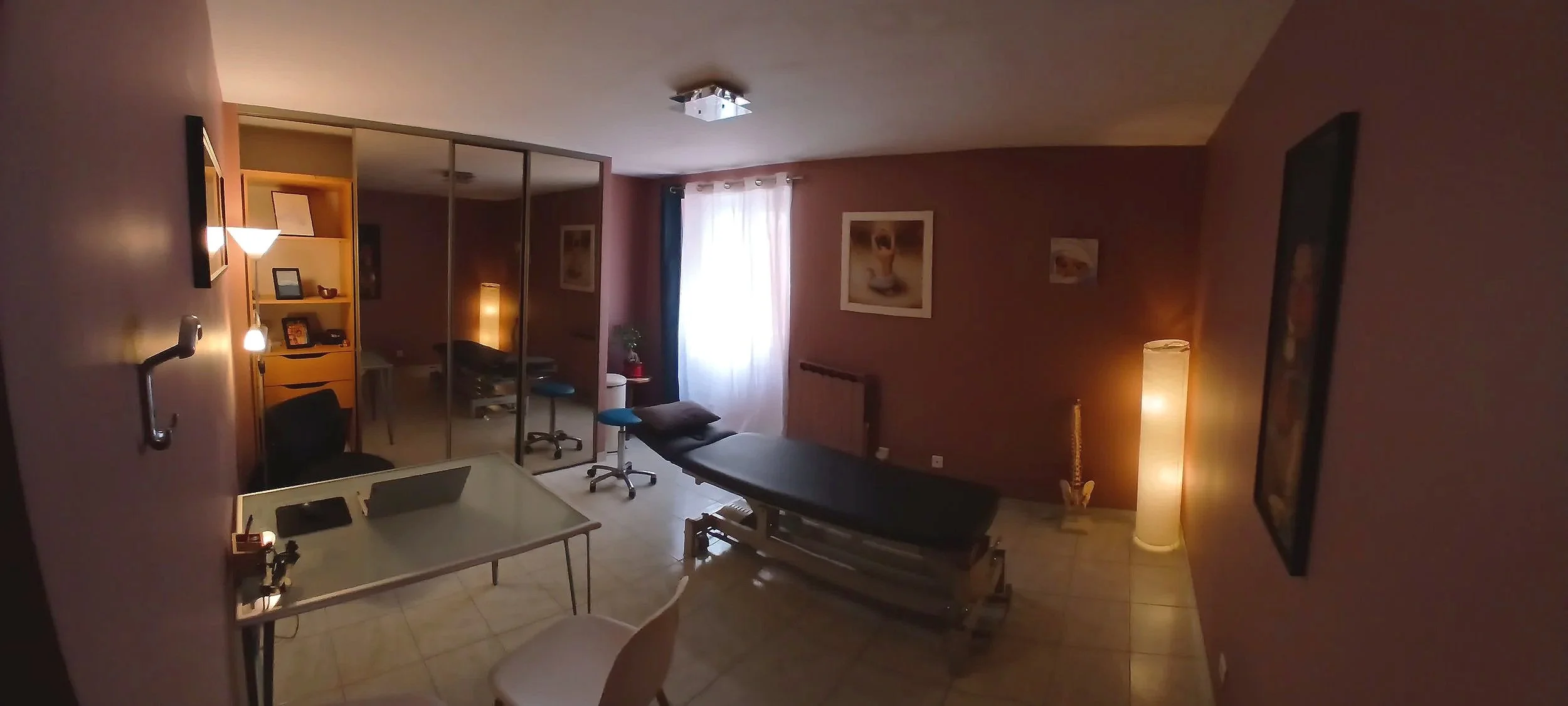 Salle de soins avec un lit d'examen, une lampe de bureau, une étagère avec des cadres, des décorations, et des lampes d'ambiance, mur rose avec des tableaux.