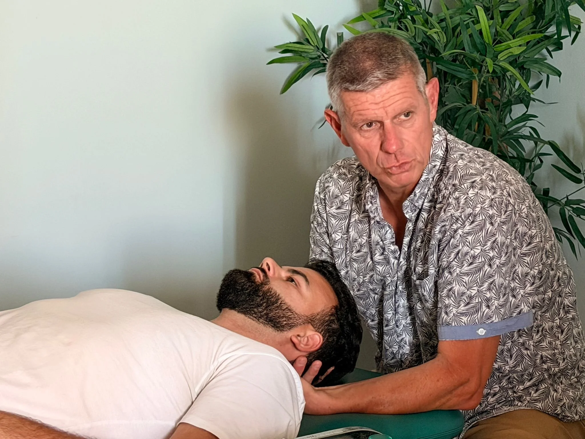 Deux hommes, dont l'un est allongé sur un lit ou une table, et un autre debout à côté, ayant une interaction avec lui, dans une pièce avec un mur blanc et une grande plante verte en arrière-plan.