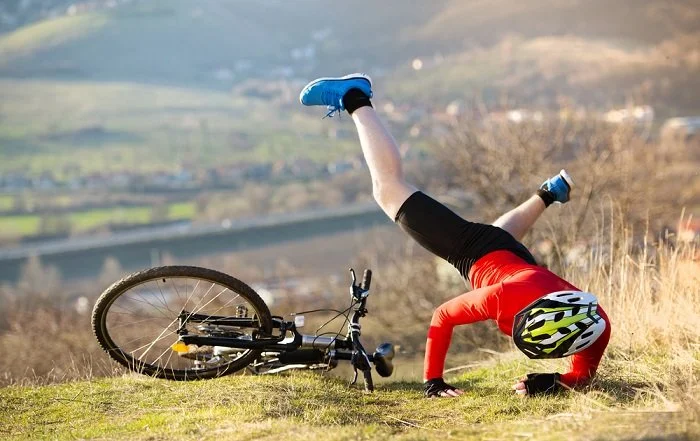Une personne en tenue de vélo effectue un saut ou une chute, avec son vélo à l'envers sur le côté, dans un paysage de campagne vallonnée.