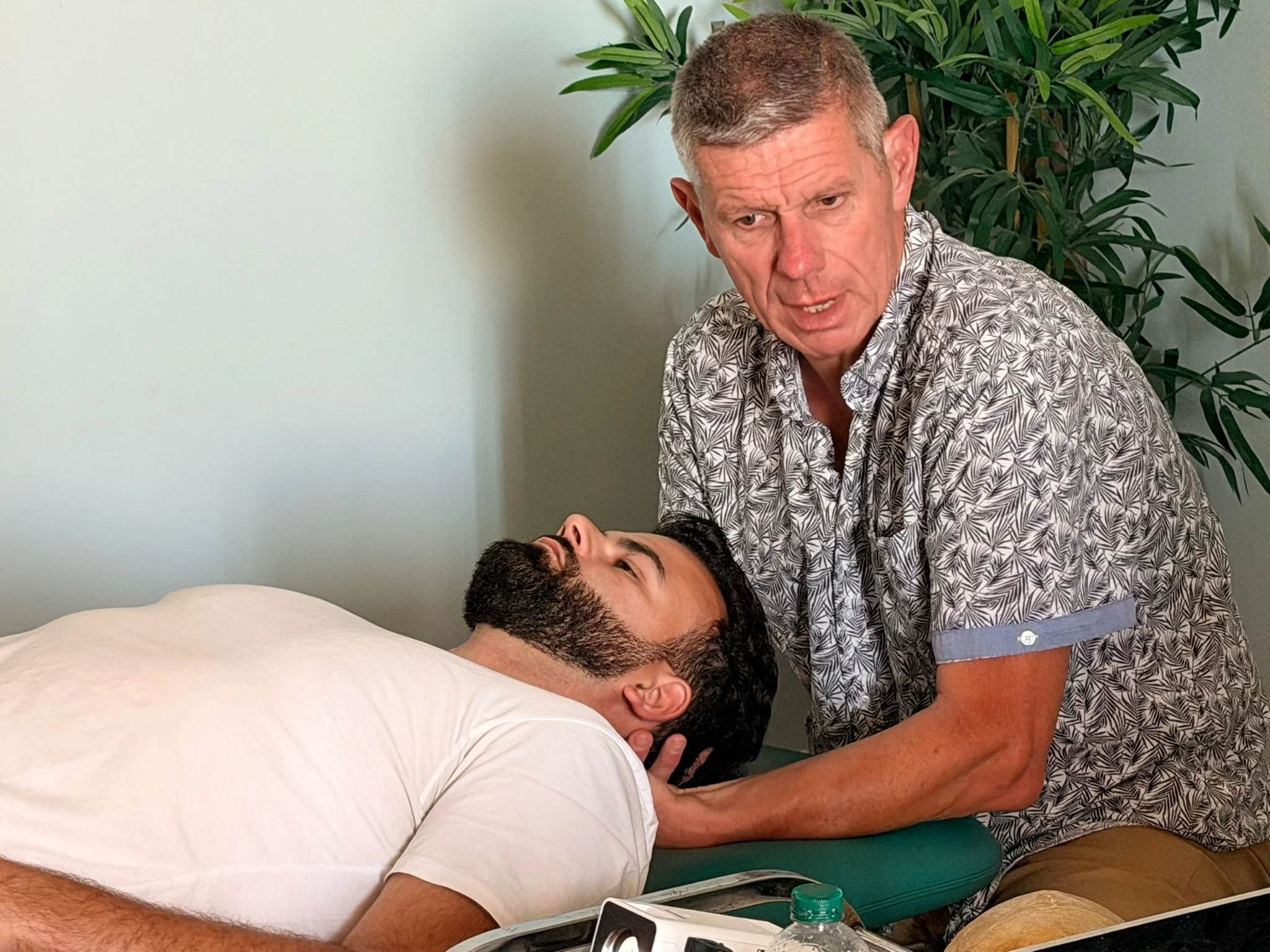 Un homme adulte allongé sur un lit, une personne se tenant au-dessus de lui, dans une pièce avec un mur blanc et une grande plante en arrière-plan.