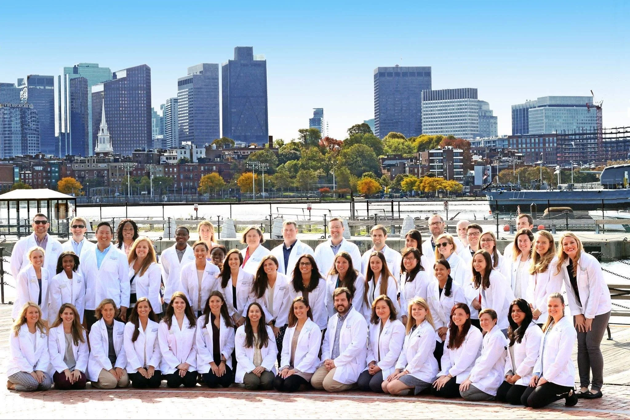A group of Physician Associates in white coats posing outdoors with the Boston city skyline in the background.