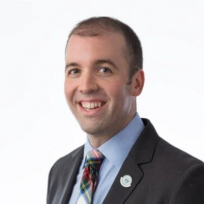 A photo of Joshua R. Merson, smiling in a suit with a blue shirt and a plaid tie, wearing a circular pin on his lapel, against a white background.