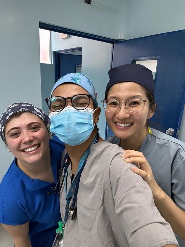 Three healthcare workers, two women and one man, smiling and taking a selfie in a medical facility. They are wearing scrubs, nurse caps, and one is wearing a mask and stethoscope.