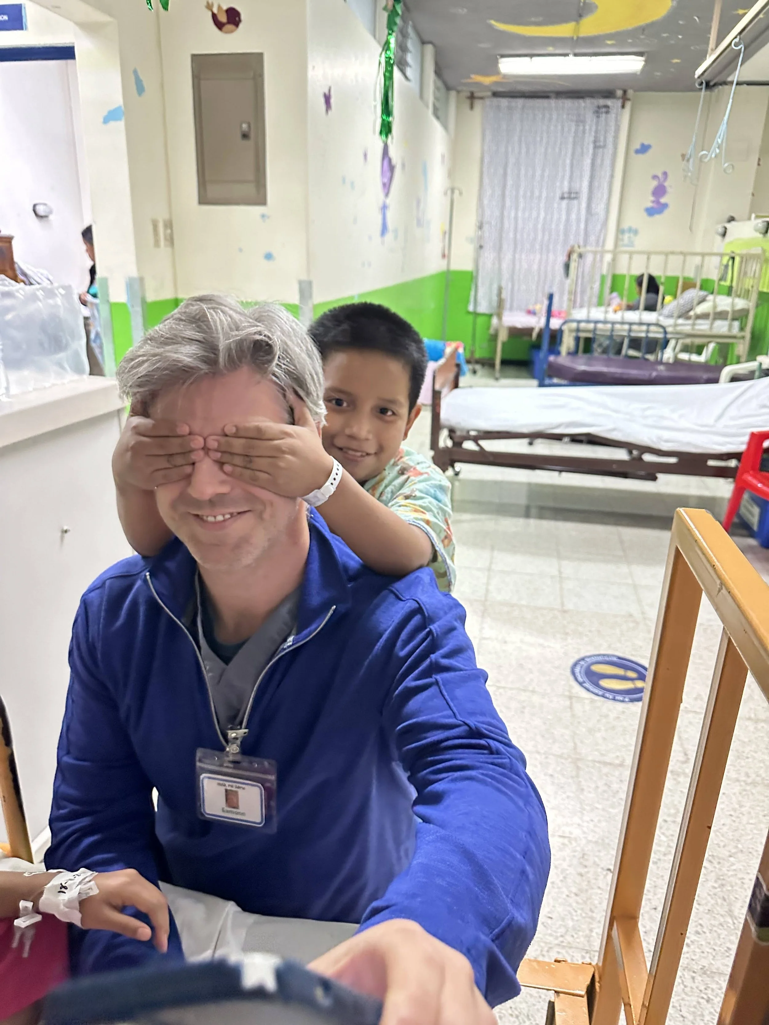 A young boy and an older man, a healthcare worker, are smiling for the camera. The boy is playfully covering the man's eyes in a hospital room with multiple beds. The room has colorful decorations on the walls.