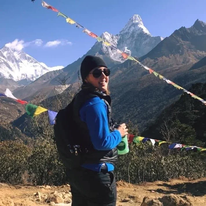 A photo of Megan Rand in outdoor gear, sunglasses, and a beanie, standing on a trail with mountains and snow-capped peaks in the background, surrounded by colorful prayer flags.