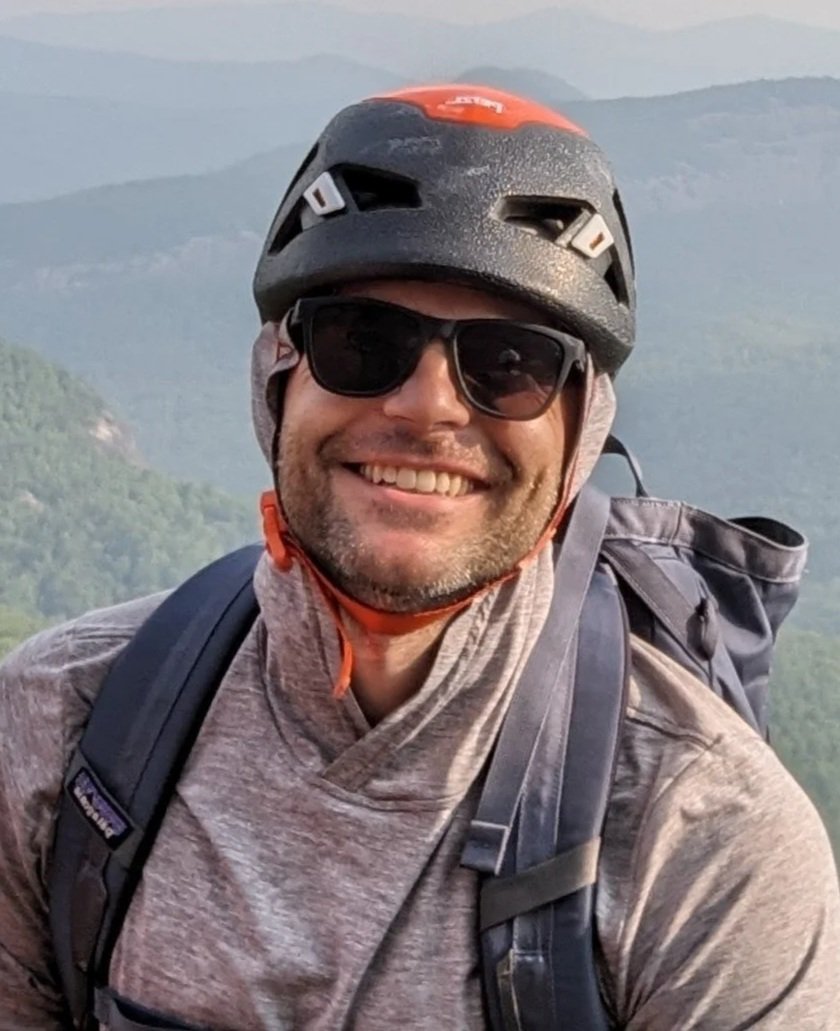 A photo of Dan McGuire wearing a black and orange bike helmet, sunglasses, and a gray jacket with a backpack, smiling outdoors with mountains and trees in the background.