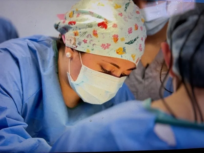 A focused photograph of a woman wearing scrubs and PPE providing care to a patient lying down while surrounded by colleagues.