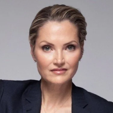 A professional portrait of Jenny Smith with short blonde hair, wearing a dark blazer, looking directly at the camera against a light gray background.