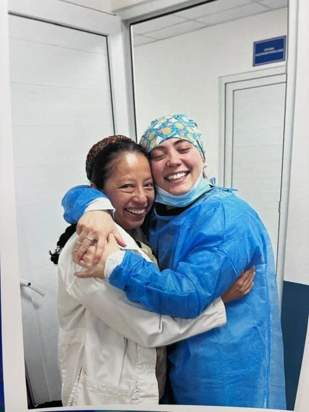 Two women, one in a doctor's coat and the other in surgical scrubs and cap, hugging and smiling in a bright clinical setting.