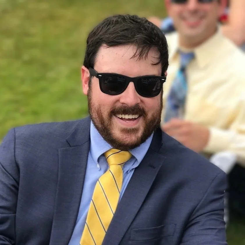 A photo of Harrison Keyes with dark hair, beard, and sunglasses wearing a dark suit, white shirt, and yellow striped tie, smiling outdoors near a body of water.