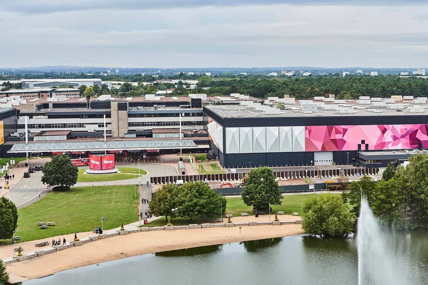 Aerial view of a large modern building with colorful geometric mural, a green park area with trees and a pond, and a fountain in the foreground. People are walking nearby.