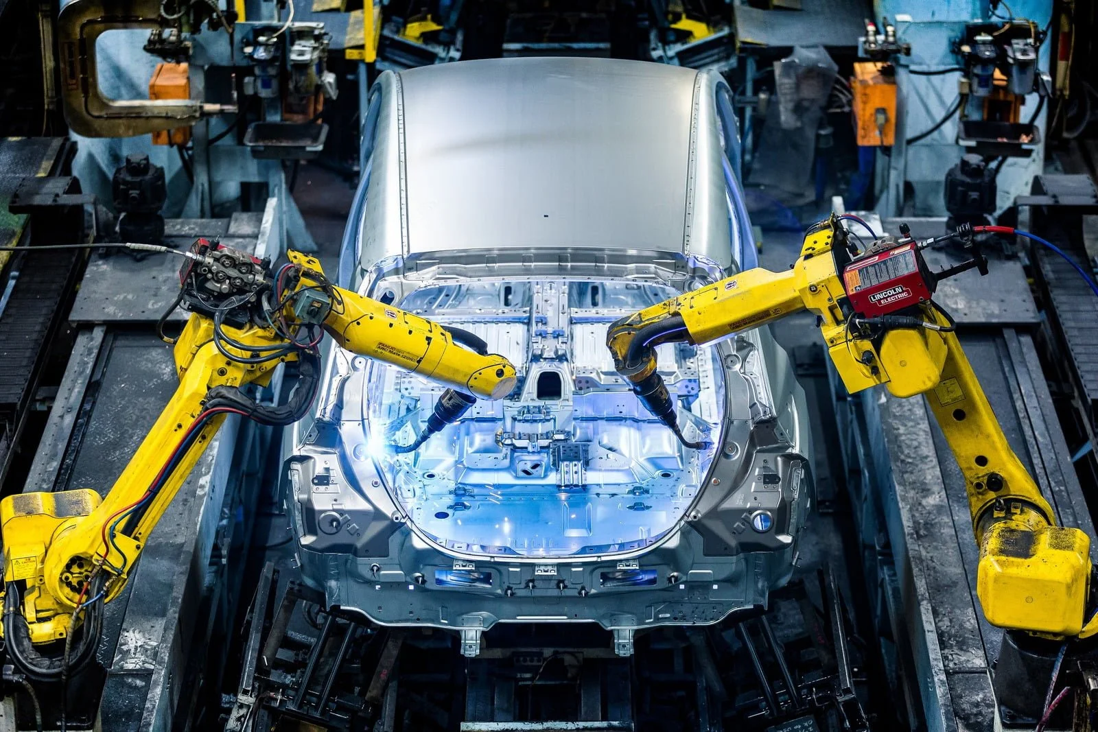 Two yellow robotic arms welding the body of a silver car in an automotive manufacturing plant.