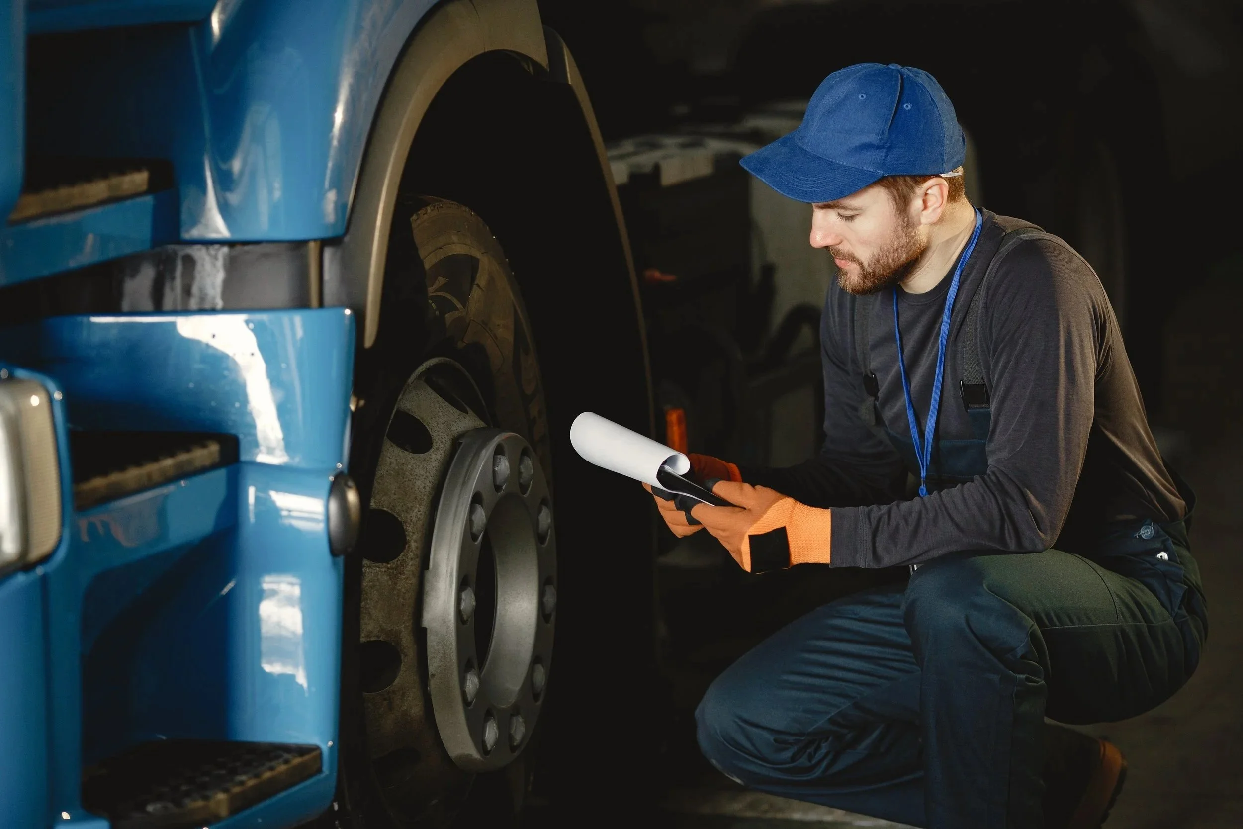 A man wearing a blue cap, black long sleeve shirt, denim pants, and orange gloves kneels beside a large blue vehicle, using a handheld device to inspect or diagnose the vehicle's front wheel area inside a dark garage or workshop.