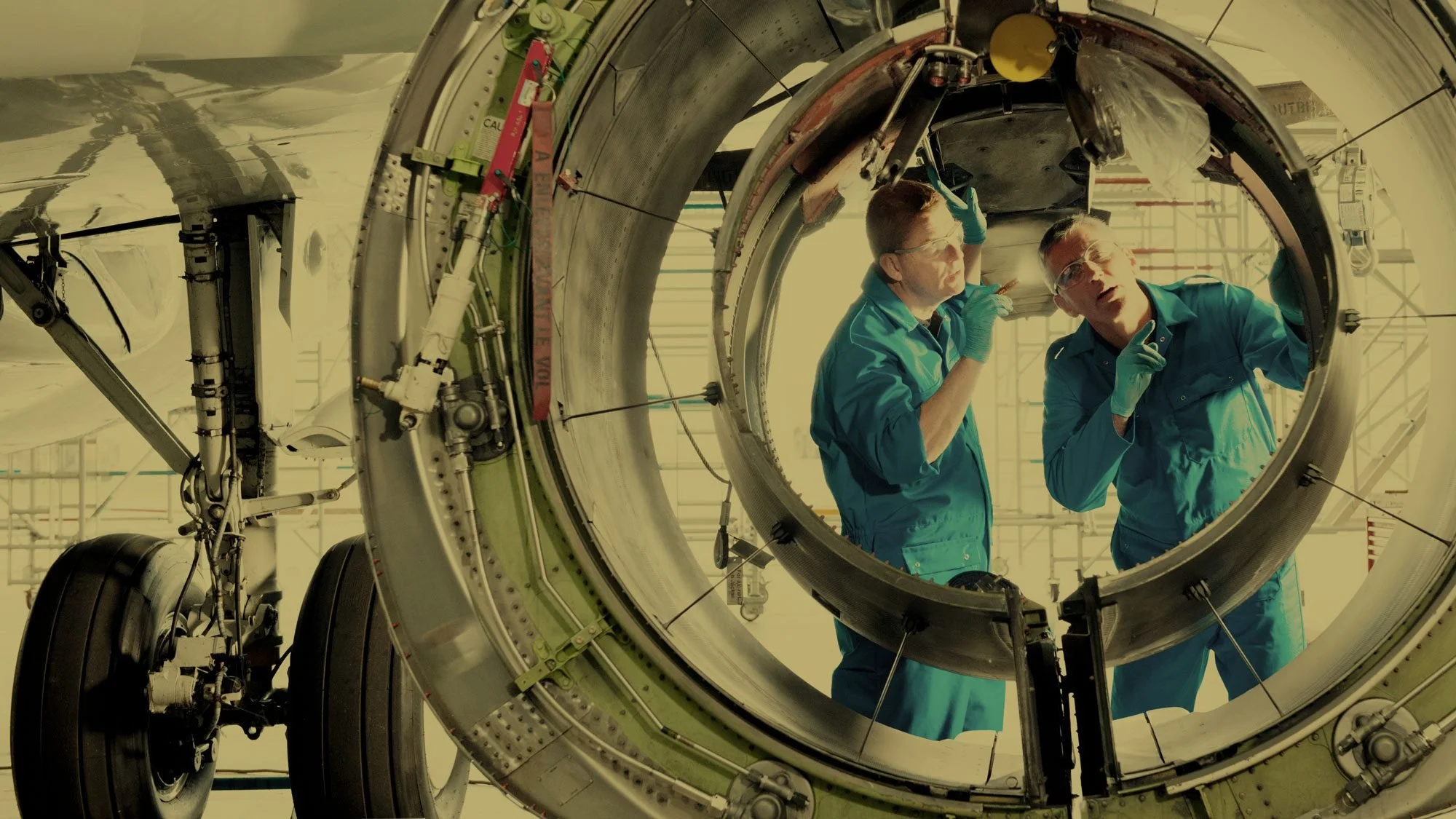 Two scientists in blue lab coats working inside a large, cylindrical spacecraft component with wheels and complex machinery at a space facility.