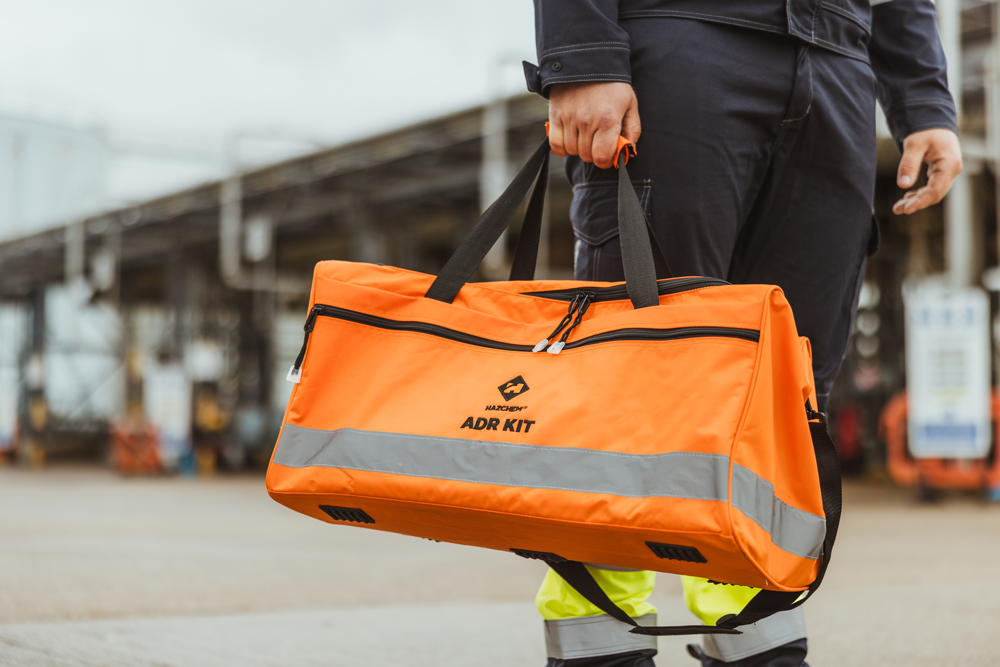 Person holding an orange emergency rescue kit bag marked 'ADR KIT' with reflective stripe, standing outdoors near an industrial area.