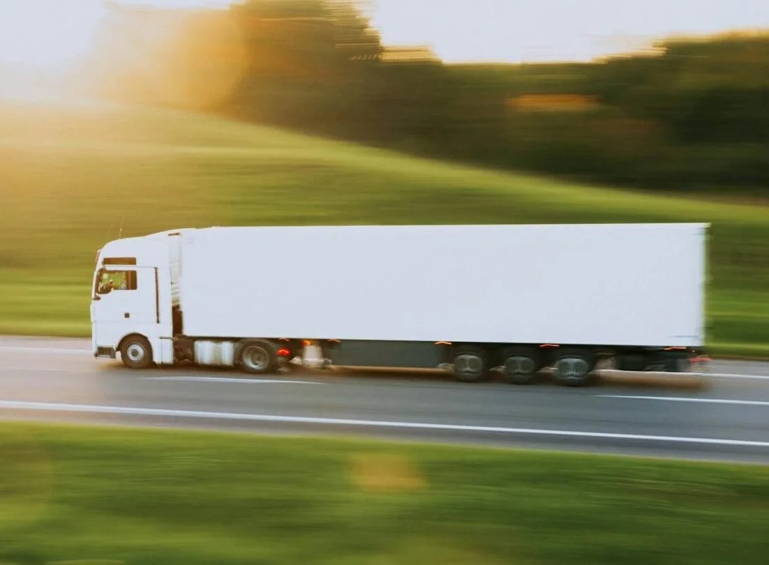 A white semi-truck driving on a highway with a blurred background of trees and sunset