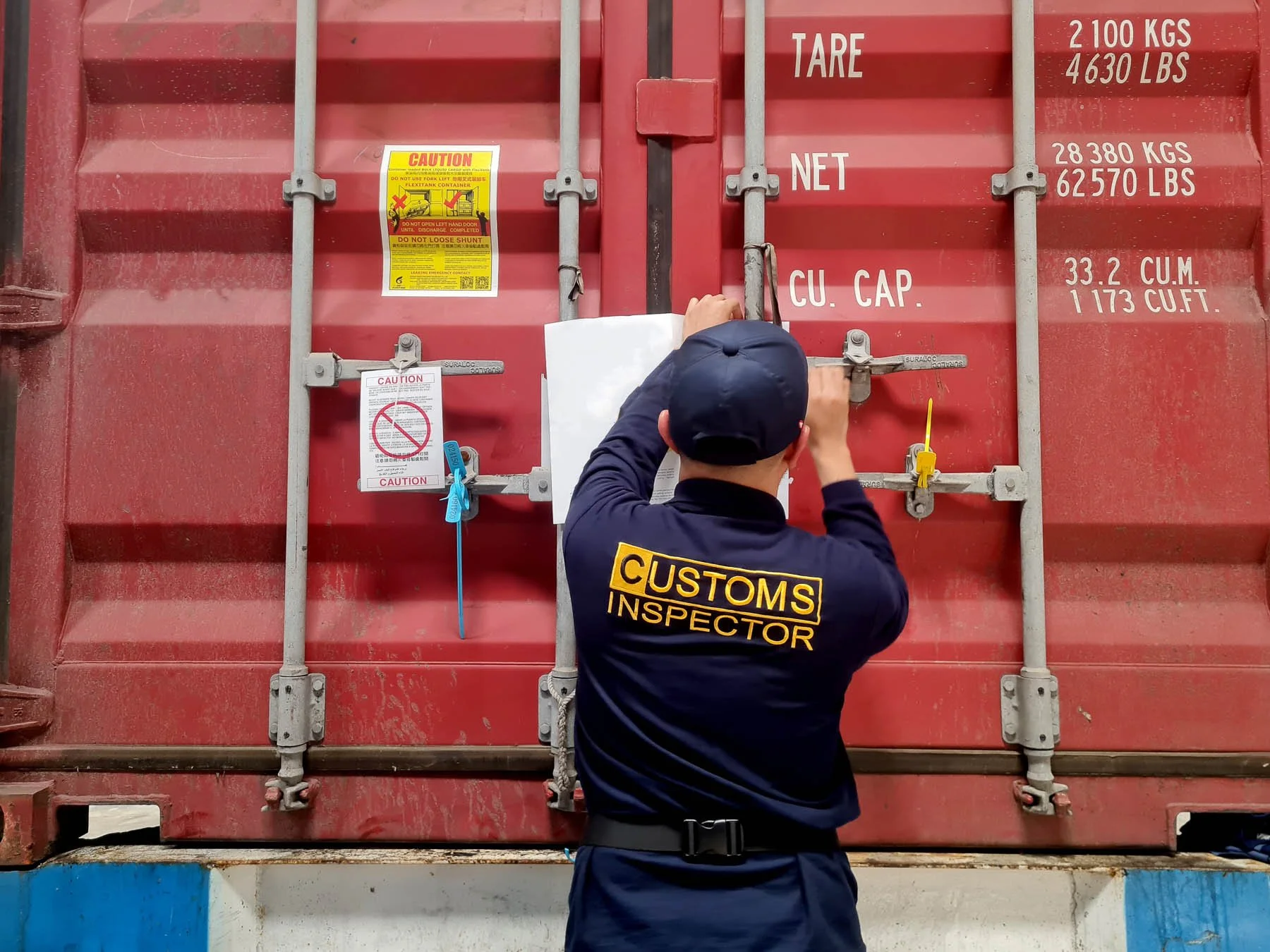 A customs inspector examines a large red shipping container. The inspector is wearing a navy uniform with a badge reading 'CUSTOMS INSPECTOR' on the back. The container has various labels and warnings, including weight limits and caution notices.