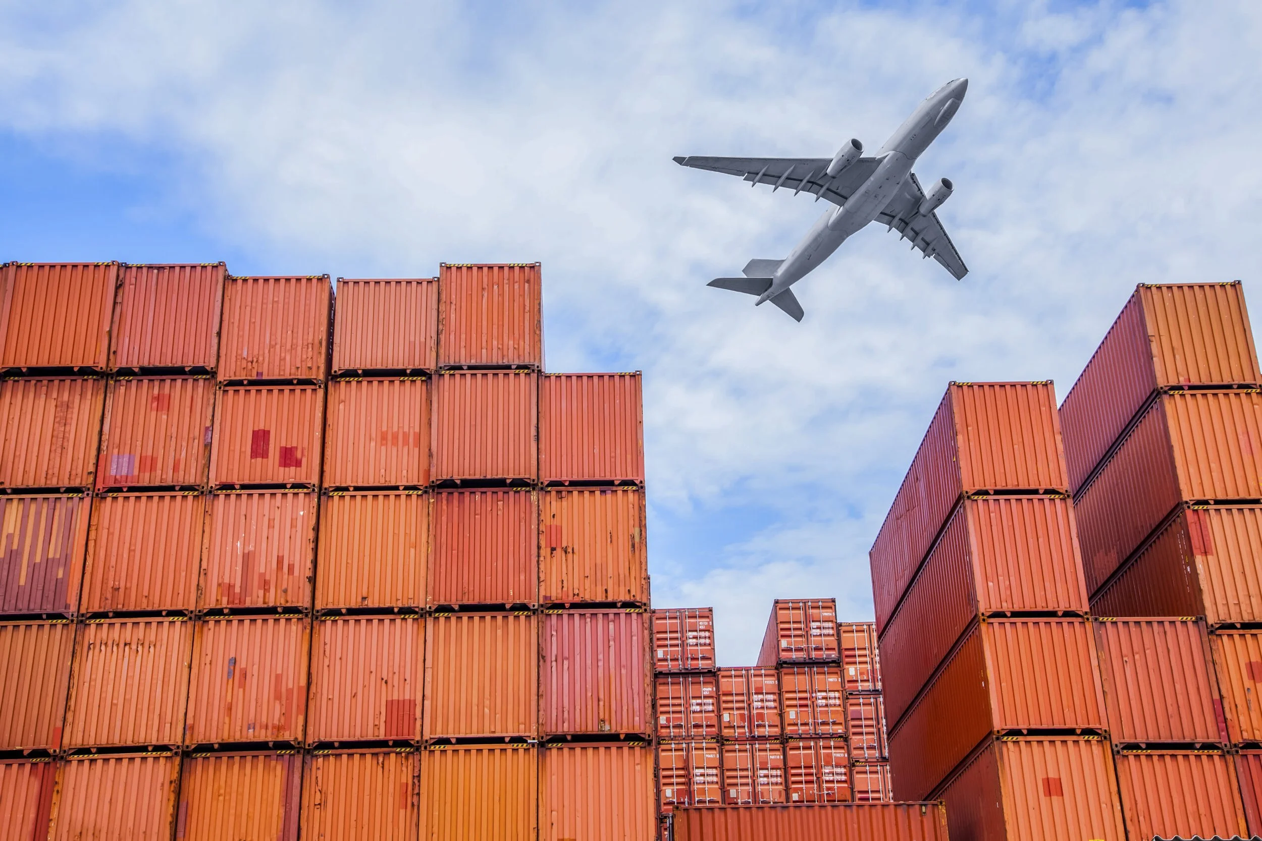 An airplane flying in the sky above stacked orange shipping containers during the daytime.