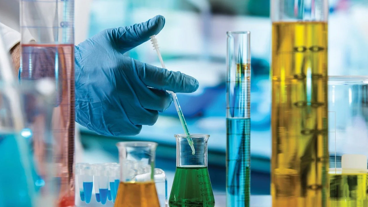 A scientist wearing blue gloves using a dropper to transfer green liquid into a beaker in a laboratory with various colorful glassware around.