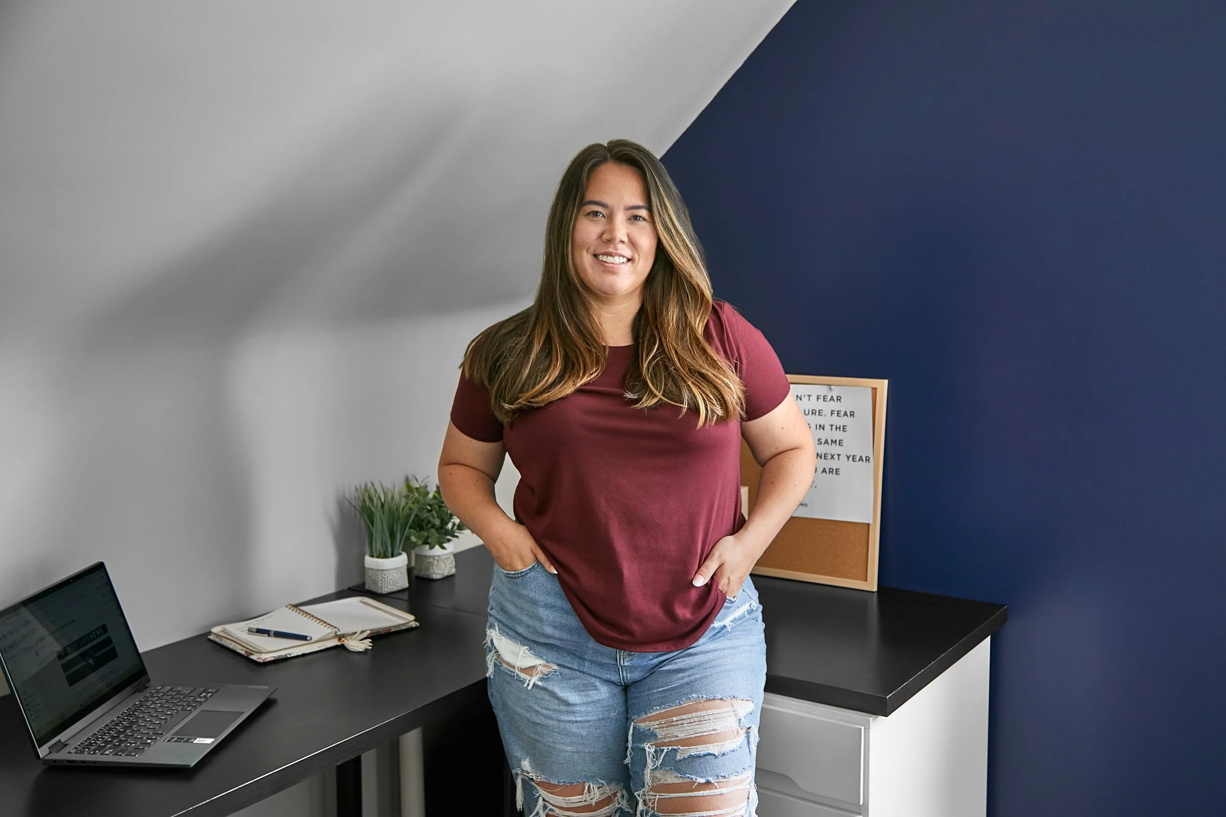 A young woman with long brown hair wearing a maroon t-shirt and ripped jeans stands in a modern office, smiling at the camera. Behind her is a desk with a laptop, notebooks, and pen, a potted plant, and a bulletin board with a motivational quote. The background wall is half gray and half dark blue.