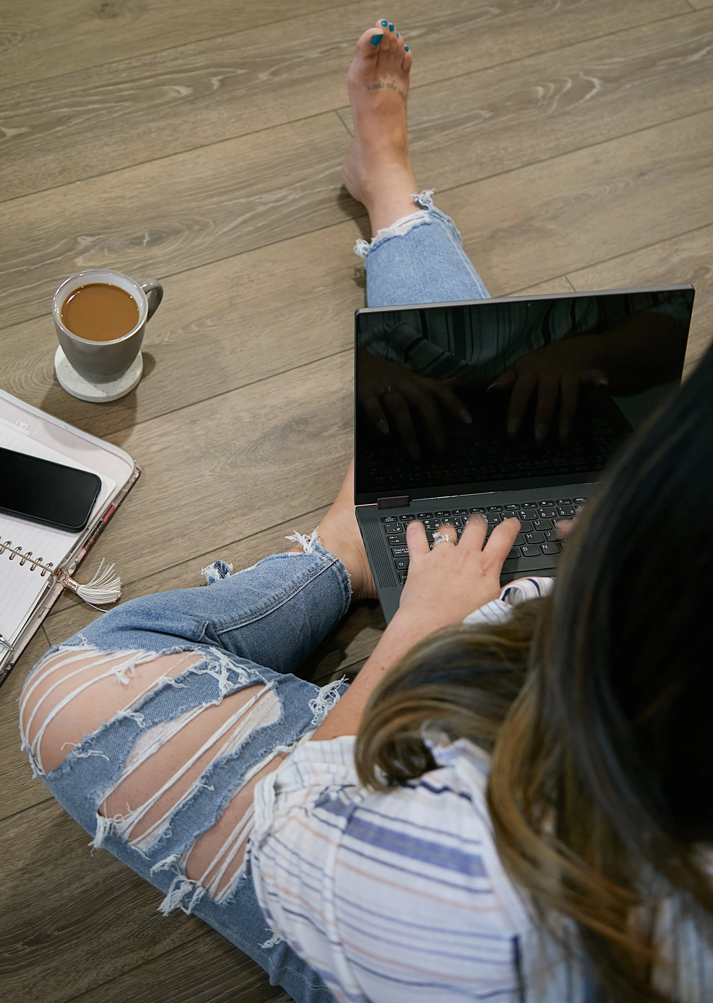 A woman is sitting on a wooden floor, wearing ripped denim shorts and a striped shirt, using a laptop with one hand and a cup of coffee nearby. She has long, wavy hair with blonde highlights.