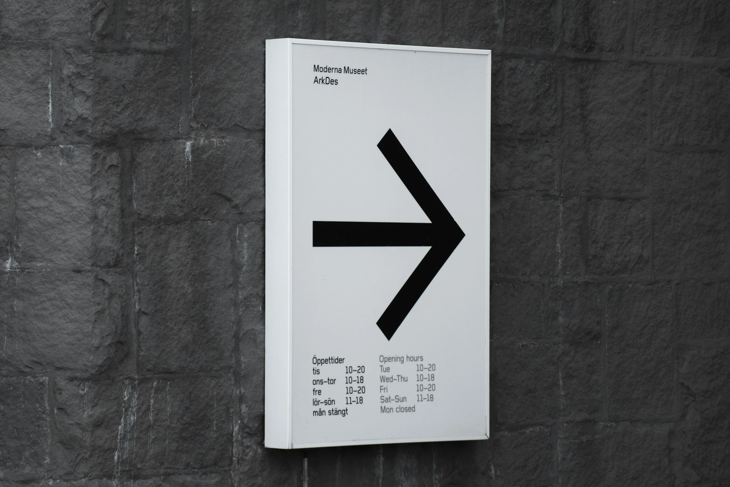 A white sign with black text and an arrow pointing right, mounted on a dark gray stone wall. The sign displays the opening hours for the Moderna Museet at ArkDes in Swedish and English, indicating it is closed on Mondays.